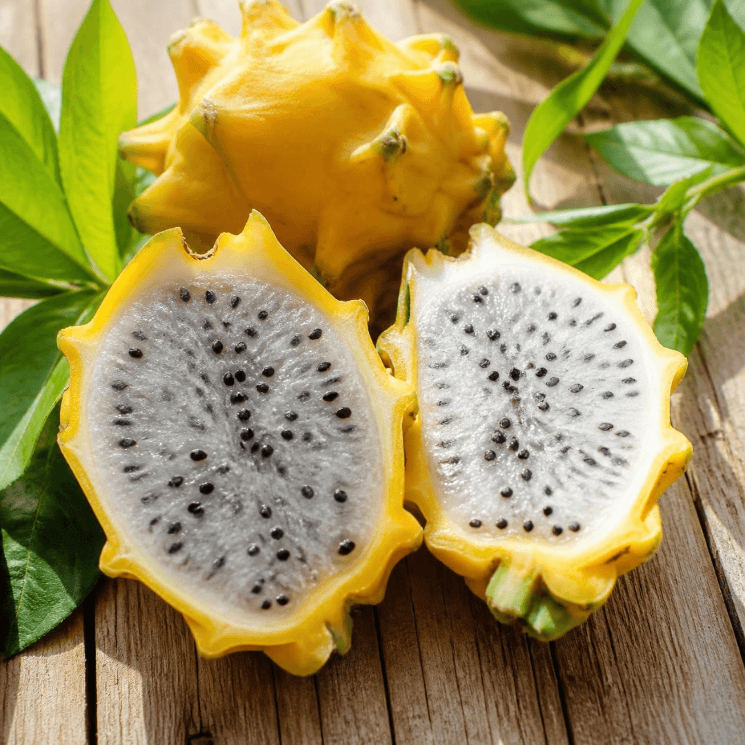 Yellow dragon fruit with one whole fruit and two halves cut open, showing white flesh with black seeds, arranged on a wooden surface with green leaves.