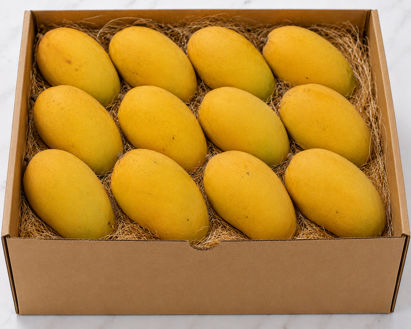 A cardboard gift box filled with ten golden dasheri mangoes resting on a bed of brown shredded paper, photographed from above on a light marble-like surface.