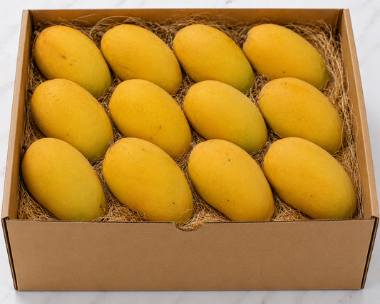 A cardboard gift box filled with ten golden dasheri mangoes resting on a bed of brown shredded paper, photographed from above on a light marble-like surface.